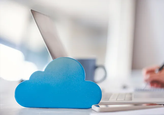 A person writing on a laptop with a cloud next to the laptop to represent cloud computing.