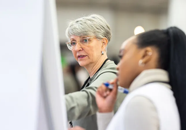 Two women stand before a flipchart and discuss it what's written on it.